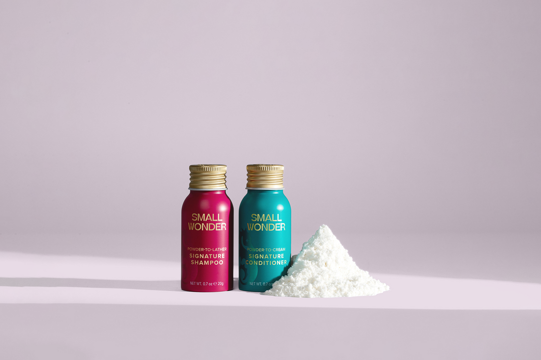 Two small, vibrant teal and magenta bottles of Small Wonder Signature Shampoo and Conditioner stand next to a neat mound of white concentrated powder on a light pink surface