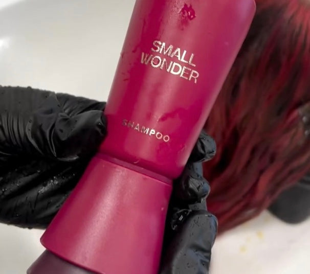 Gloved professional hands holding red Small Wonder shampoo bottle above customer's hair
